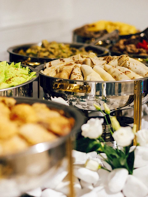 A buffet setup with various dishes in silver chafing dishes, surrounded by elegant white decorations and flowers.