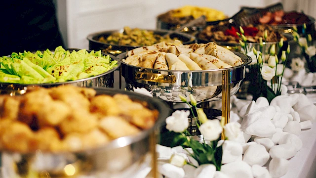A buffet setup with various dishes in silver chafing dishes, surrounded by elegant white decorations and flowers.