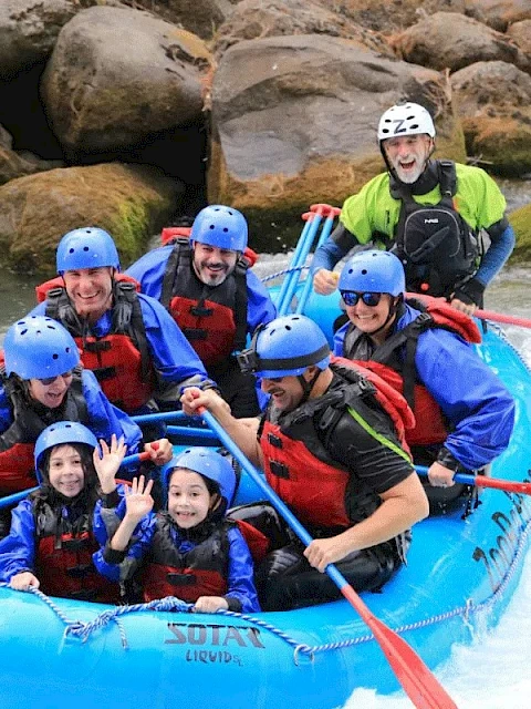 A group of people wearing helmets and life jackets are white-water rafting in a blue inflatable raft, smiling and waving.