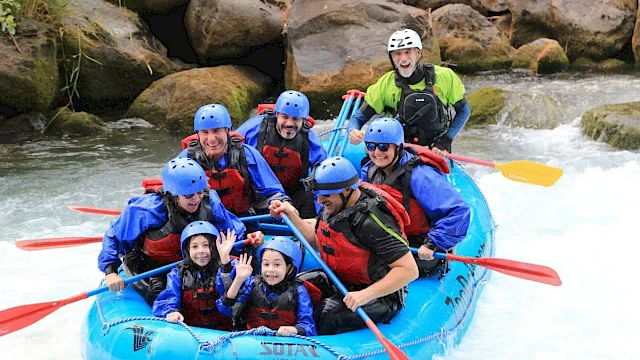 A group of people wearing helmets and life jackets are white-water rafting in a blue inflatable raft, smiling and waving.