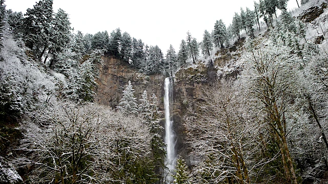 A tall waterfall in a cliffside forest, light snow on trees, misty spray at the base, wintry scene with evergreen pines.