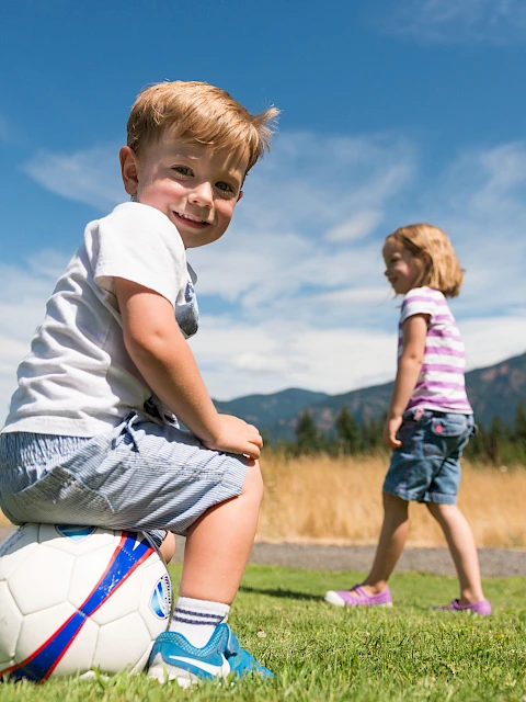 Two young children play soccer on a sunny field, with one boy sitting on a ball in the foreground as another girl jogs in the background.