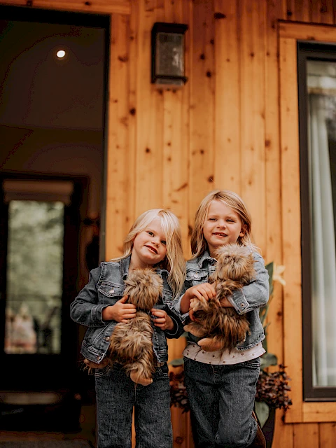 Two children in denim jackets stand outside a wooden house, each holding a small fluffy dog, smiling.