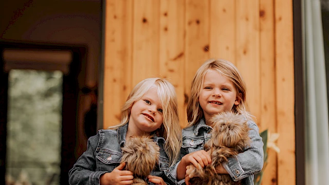 Two children in denim jackets stand outside a wooden house, each holding a small fluffy dog, smiling.