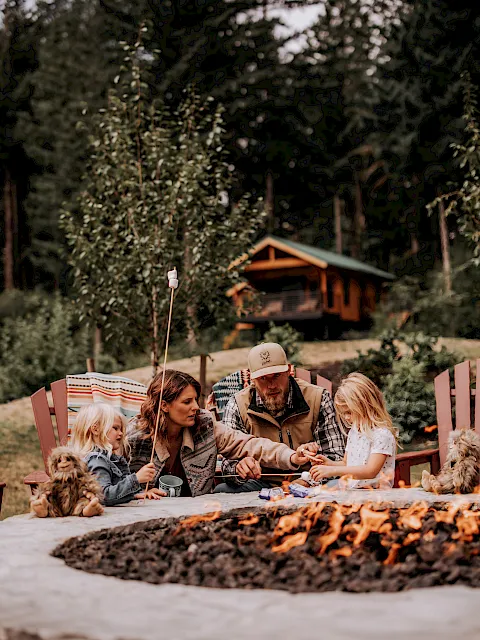 A family enjoys an outdoor campfire picnic with kids and pets in front of lush trees and a cabin, sharing warm moments together.