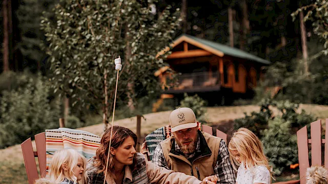 A family enjoys an outdoor campfire picnic with kids and pets in front of lush trees and a cabin, sharing warm moments together.