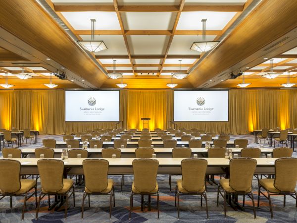 A conference room is set up with rows of tables and chairs facing two large screens displaying a logo.