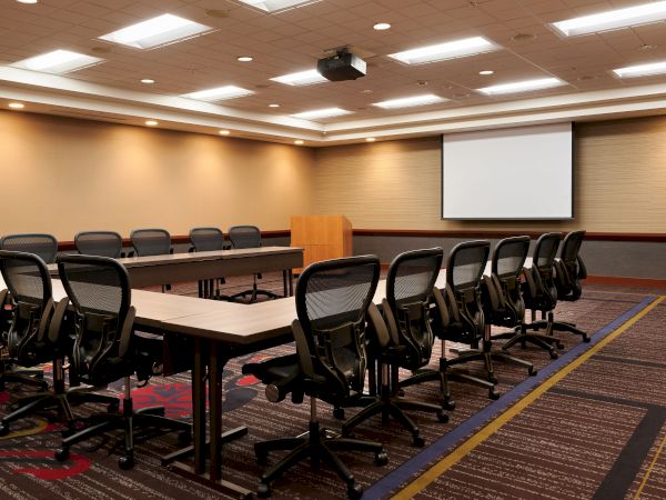 A conference room with a U-shaped table setup, office chairs, a wooden podium, ceiling lights, and a projection screen on one wall.