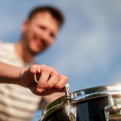 A person is adjusting a drum's tension rod, focusing on tuning the instrument, against a blurred outdoor background.