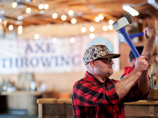 A person in a plaid shirt and cap is preparing to throw an axe in an indoor axe-throwing venue.