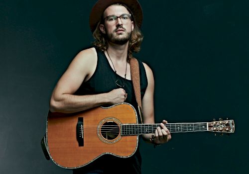 A person wearing a hat and glasses stands against a dark background, holding an acoustic guitar.