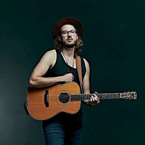 A person wearing a hat and glasses stands against a dark background, holding an acoustic guitar.