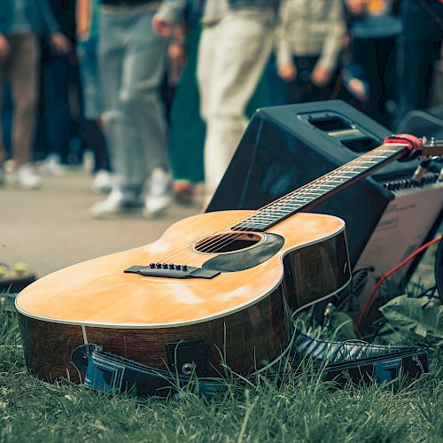 An acoustic guitar lies on the grass near a microphone, speakers, and an effects pedal, with a blurred crowd in the background.