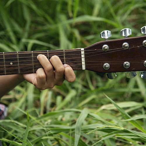 A person playing an acoustic guitar in a field of green grass, with their hand on the fretboard.