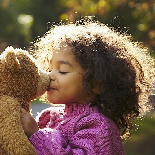 A child in a pink sweater kisses a teddy bear outdoors in a sunlit setting.