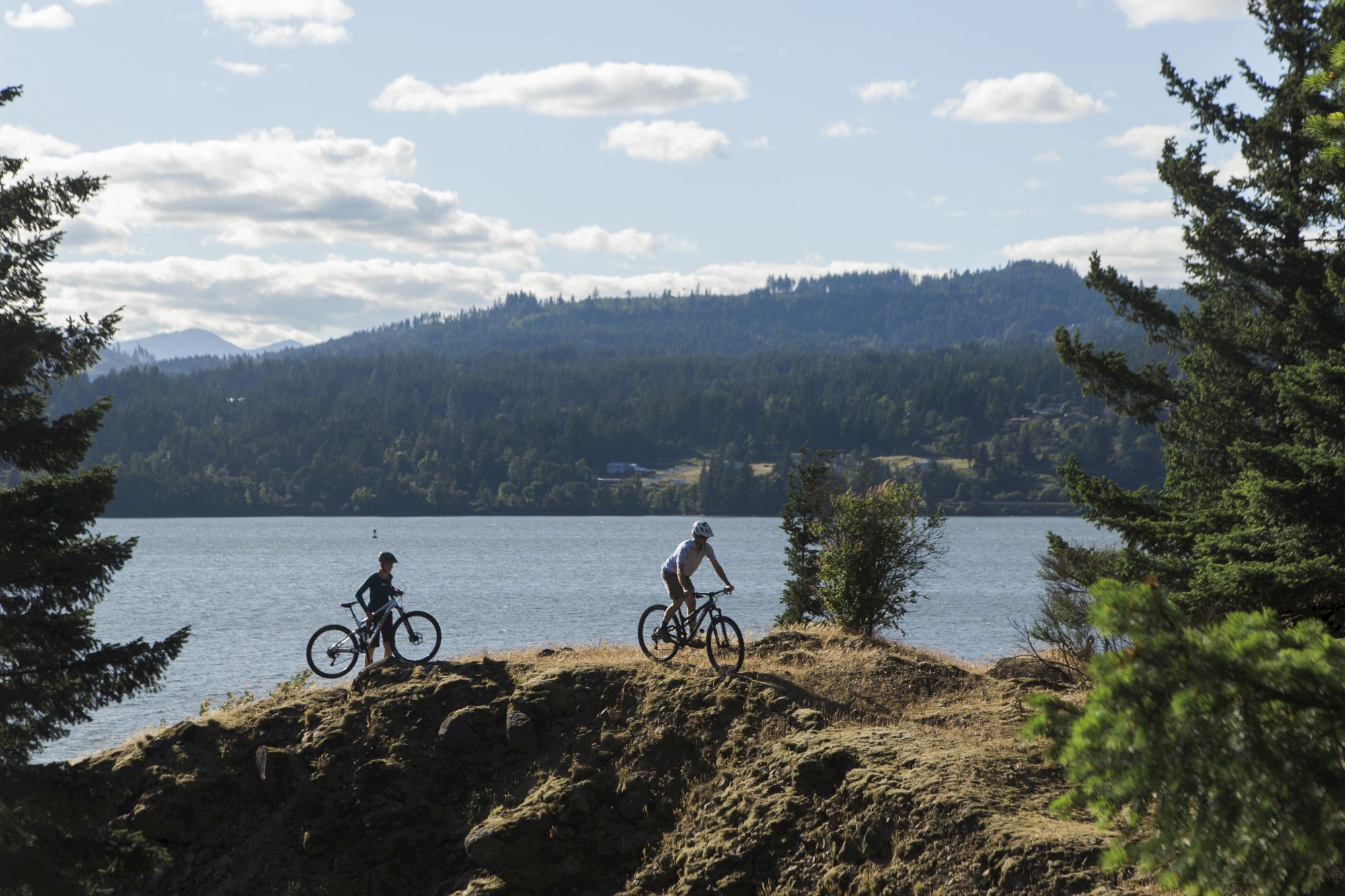 Two people are biking along a scenic lakeside trail with mountains and trees in the background under a partly cloudy sky.