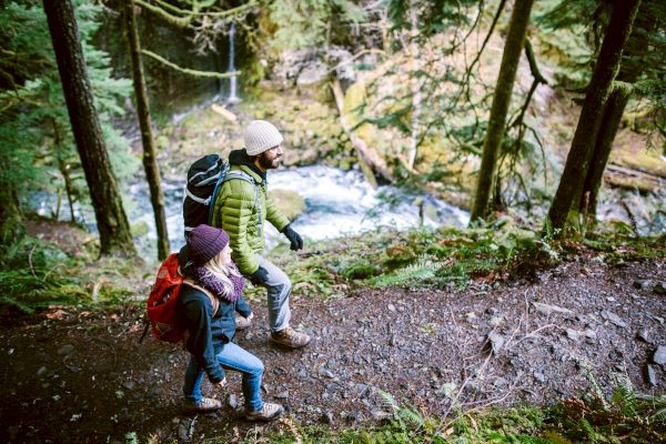 Two people hiking on a forest trail with backpacks, enjoying the outdoors near a stream.