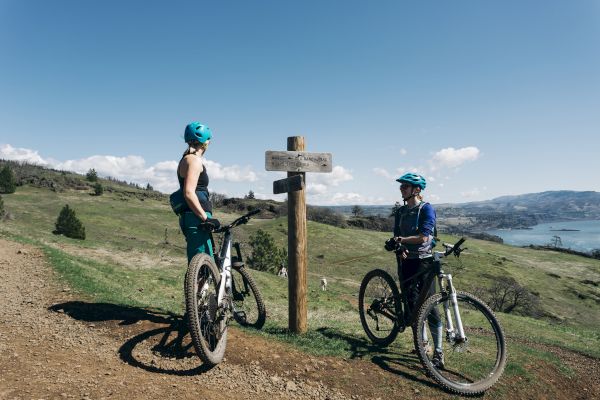 Two cyclists in helmets, stopped at a signpost on a grassy trail, overlook a scenic landscape with hills and a body of water.