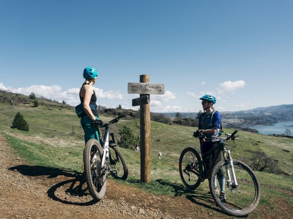 Two cyclists in helmets, stopped at a signpost on a grassy trail, overlook a scenic landscape with hills and a body of water.