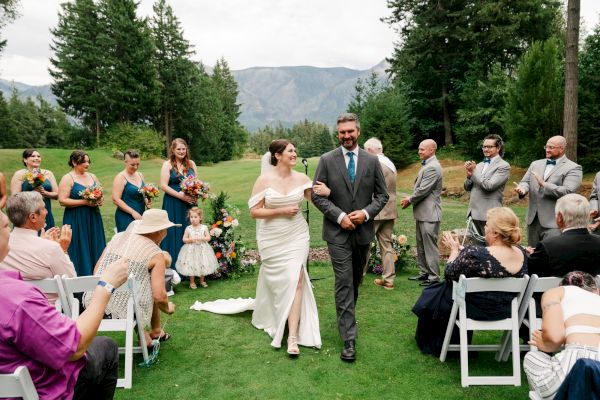 A couple walks arm in arm down an outdoor aisle, surrounded by happy guests and a wedding party, while people applaud in celebration.