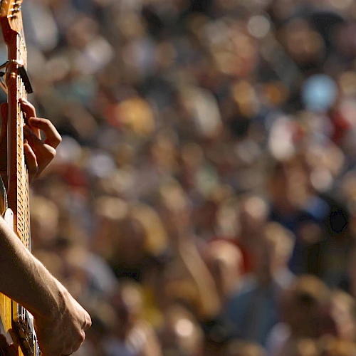 A person is playing a guitar in the foreground, with a blurred crowd in the background, possibly at an outdoor concert or festival.