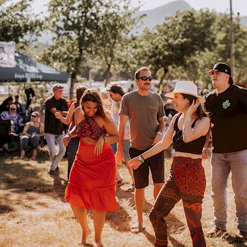 People are dancing outdoors at a lively gathering, with sunlight and trees in the background.