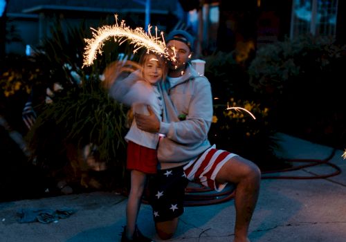 A person kneels, holding a young child with a sparkler making light trails in the air, evoking a joyful and festive atmosphere.
