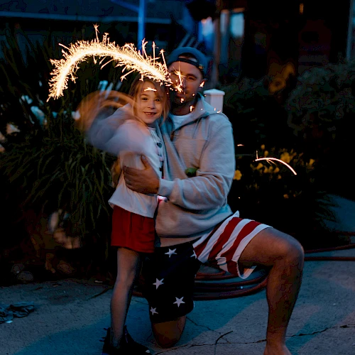 A person kneels, holding a young child with a sparkler making light trails in the air, evoking a joyful and festive atmosphere.
