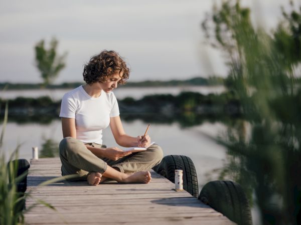 A person is sitting cross-legged on a wooden dock by the water, writing in a notebook, surrounded by nature.