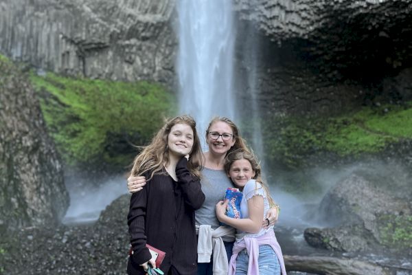 A group of three people posing happily in front of a waterfall, surrounded by lush greenery and rocky terrain.