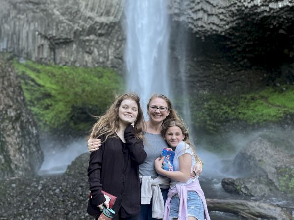 A group of three people posing happily in front of a waterfall, surrounded by lush greenery and rocky terrain.