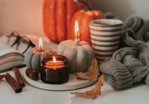 Cozy autumn scene with lit candles, pumpkins, a striped mug, knitted blanket, an open book, glasses, and cinnamon sticks on a white surface.