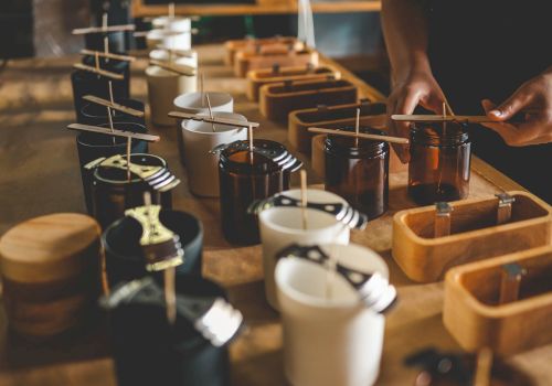 The image shows a person arranging wicks in jars for candle making, with various tools and containers on a wooden table.