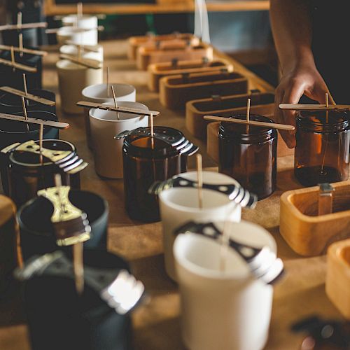 The image shows a person arranging wicks in jars for candle making, with various tools and containers on a wooden table.