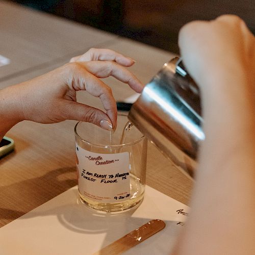 A person pours liquid into a glass while holding a straw, surrounded by papers and a knife on a table.