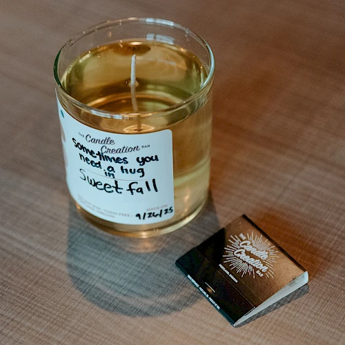 A candle labeled "Sweet Fall" with text on it, next to a matchbox, is placed on a wooden surface.