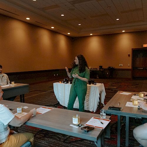 A woman is speaking to a group seated at tables in a meeting room; papers and cups are on the tables.