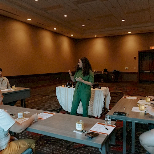A woman is speaking to a group seated at tables in a meeting room; papers and cups are on the tables.