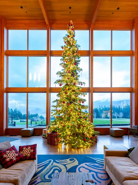 A large decorated Christmas tree with glowing lights and ornaments stands in a cozy wooden living room by snowy windows.