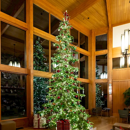 A tall Christmas tree with lights and ornaments stands in a wooden interior with large windows and cozy furniture.