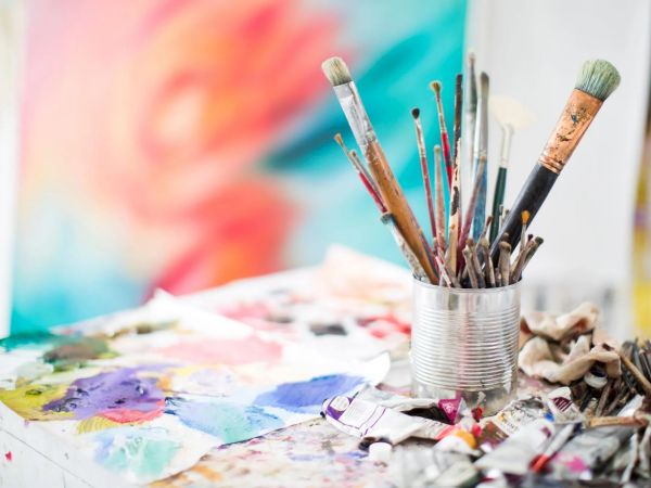 Paintbrushes in a tin can and tubes of paint are on a table with a colorful abstract painting blurred in the background.