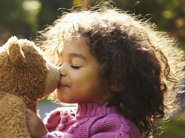 A child with curly hair kisses a teddy bear while wearing a purple sweater in a sunny outdoor setting.