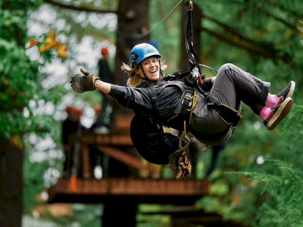 A person wearing a helmet and safety gear is zip-lining through a forest, smiling and giving a thumbs-up, with trees in the background.