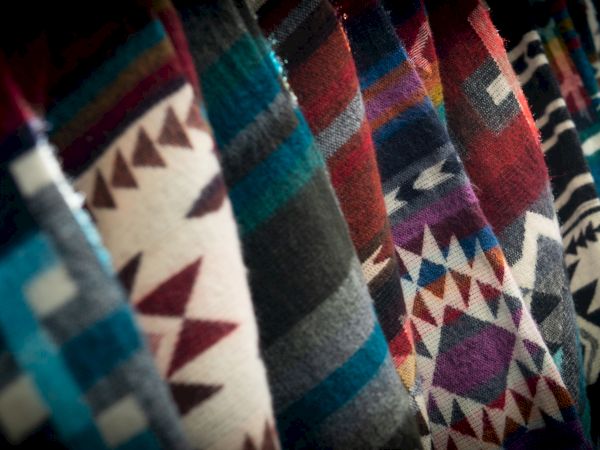 The image shows a row of colorful, patterned fabrics with geometric designs, possibly woven blankets or textiles, displayed vertically.