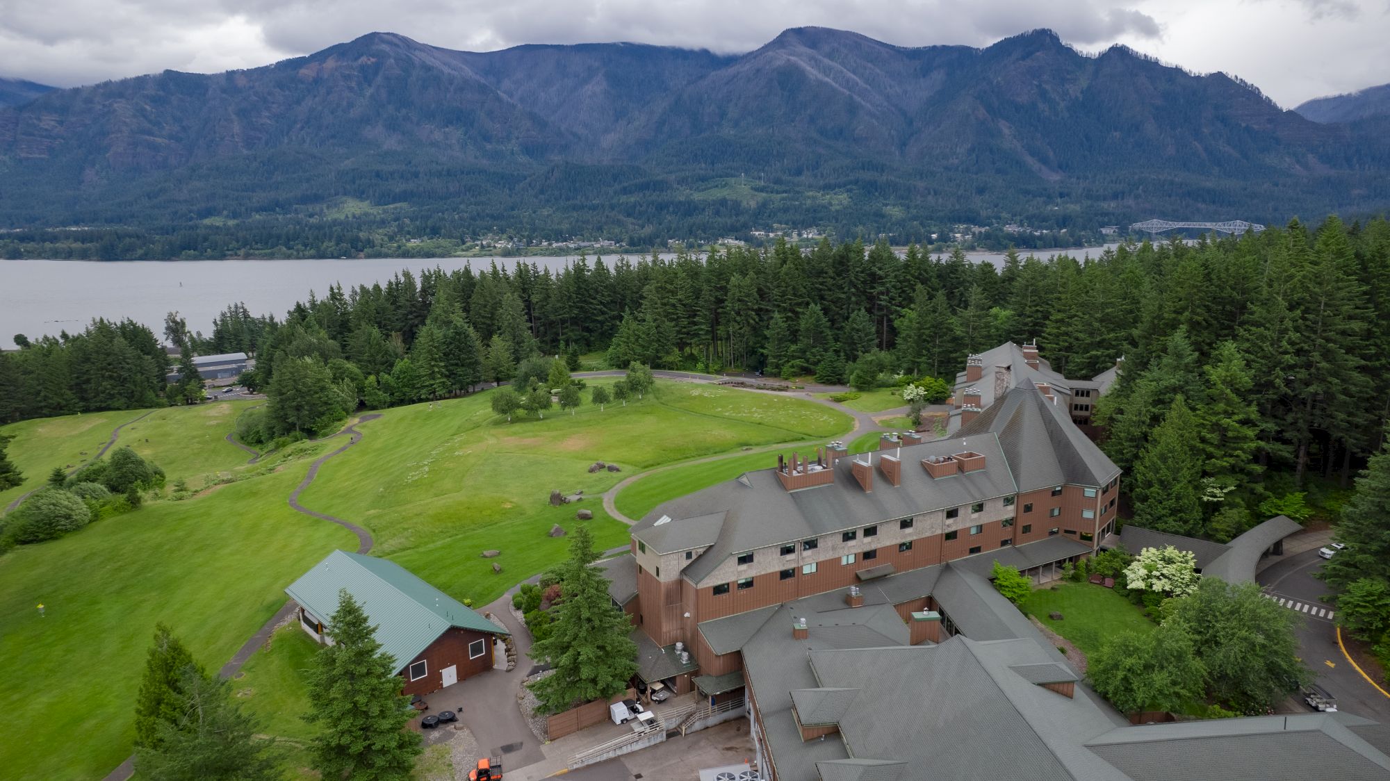 A scenic view features a large building surrounded by lush greenery, with a backdrop of mountains and a lake under a cloudy sky.