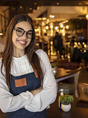 A person is smiling, standing inside a warmly lit restaurant with tables and chairs in the background, wearing glasses and an apron.