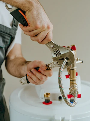 A person in overalls uses a wrench on a water heater valve, with red fittings and piping visible on top of the tank.