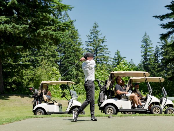 A golfer swings on a course, with two golf carts and trees in the background.