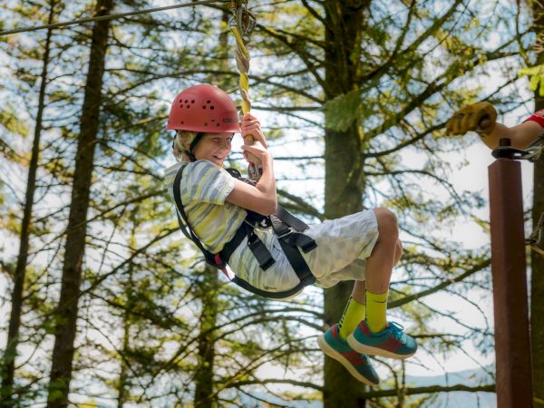 A person in a helmet and harness is enjoying a zip line ride amidst a forest, assisted by an instructor.