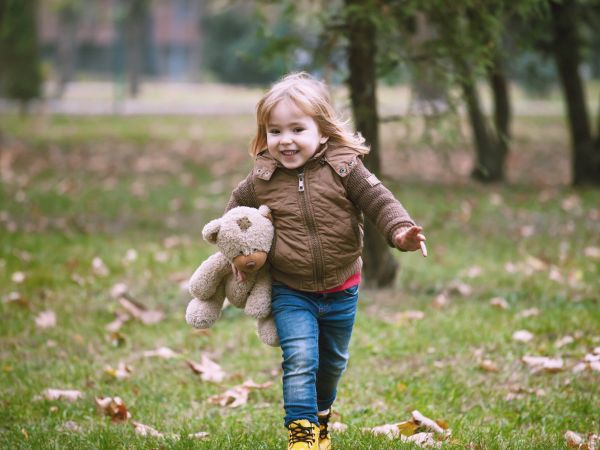 A smiling child in a brown jacket walks through a grassy area while holding a teddy bear. Leaves are scattered on the ground.